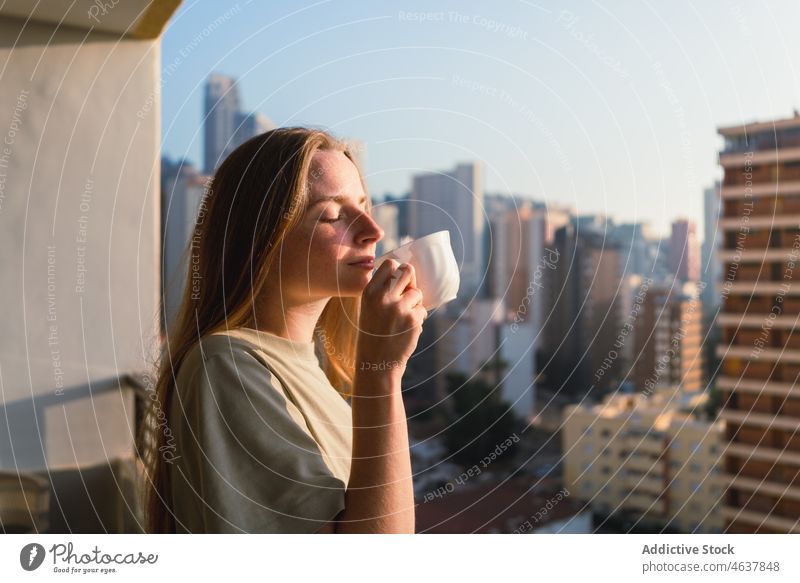Calm young woman enjoying coffee smell on balcony sunny in morning drink terrace city weekend eyes closed relax peaceful female long hair t shirt carefree calm