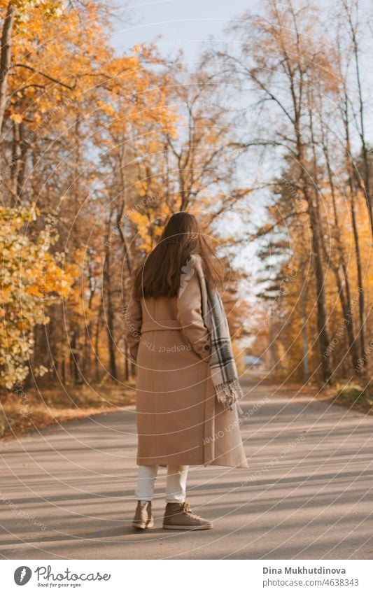 Young brunette woman with long hair in beige coat walking in autumn park on a road. Relaxing moments in nature, millennial girl being happy and smiling. Woman from behind walking in autumn park.