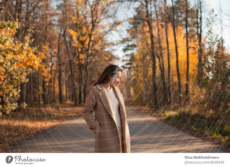 Young brunette woman with long hair in beige coat walking in autumn park on a road. Relaxing moments in nature, millennial girl being happy and smiling. Woman smiling walking in autumn park.
