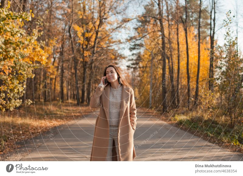 Young brunette woman with long hair in beige coat walking in autumn park on a road. Relaxing moments in nature, millennial girl being happy and smiling. Woman talking on the phone walking in autumn park.