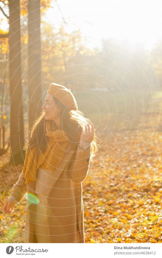Young woman in yellow beret and scarf and in beige coat walking in autumn park at sunset with warm sunshine. Relaxing moments in nature, millennial girl being happy and smiling.