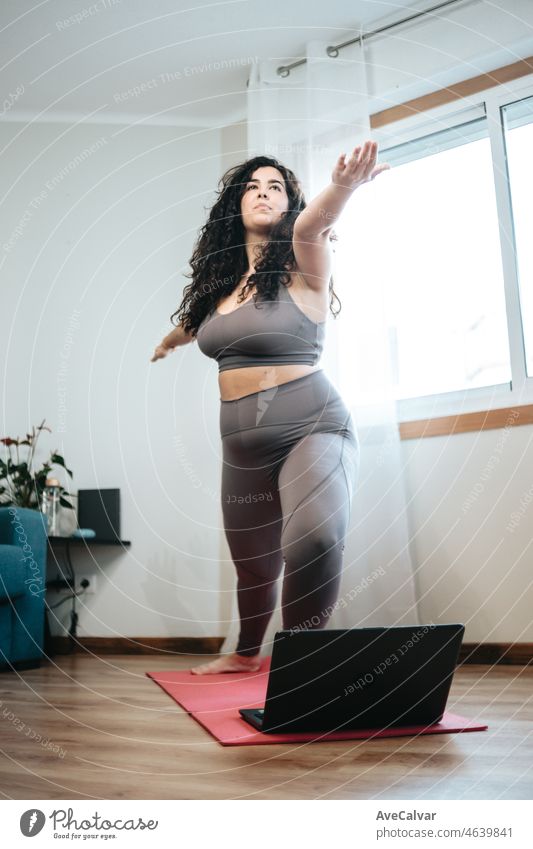 Full length shot of young plus size woman in sportswear doing yoga positions on the floor with a laptop. Copy space. Losing weight gaining health at home. Online class with the laptop concept.
