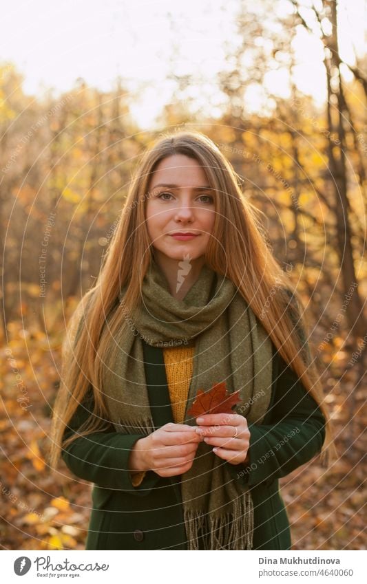 Beautiful woman with long hair in green coat standing in autumn park holding a fall maple leaf. Style, fashion and clothes. Deep in thought in autumn forest in nature. Authentic lifestyle.