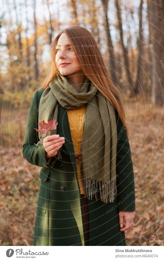 Beautiful woman with long hair in green coat standing in autumn park holding a fall maple leaf. Style, fashion and clothes. Deep in thought in autumn forest in nature. Authentic lifestyle.