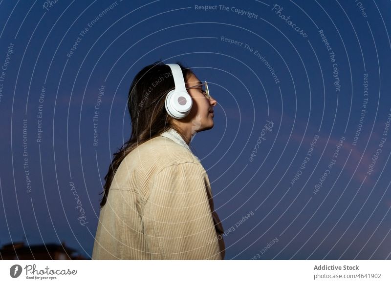 Anonymous woman listening to music in headphones at dusk wireless happy playlist sunset song teenage enjoy female delight positive device modern connection
