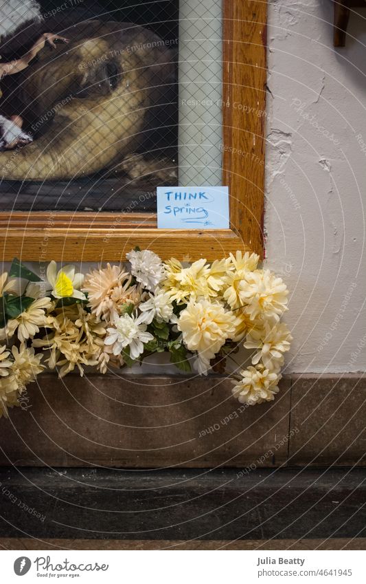 Odd combination of fake flowers, corner of a painting, and a hand written sign tht says "think spring" faux nature plastic bunch arrangement odd weird