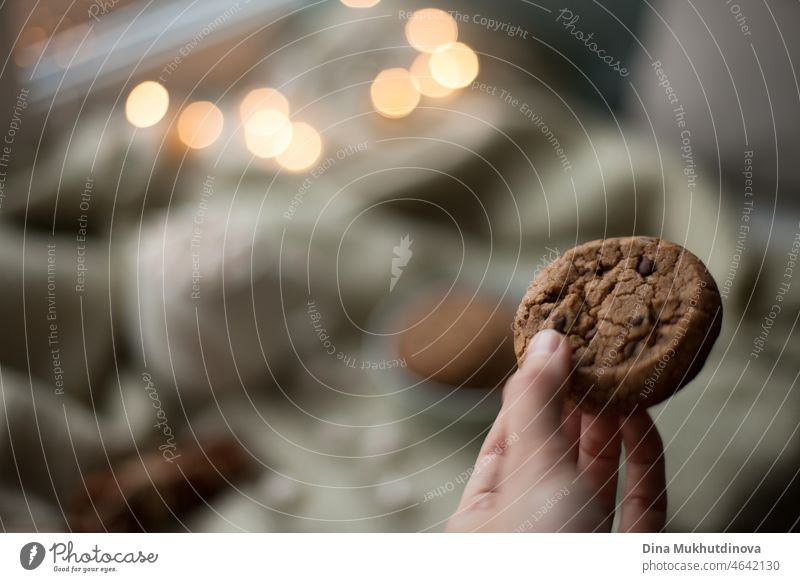 Hand holding a homemade cookie close up. Homemade chocolate chip cookies at cozy home with lights bokeh. Copy space and low key evening background food dessert