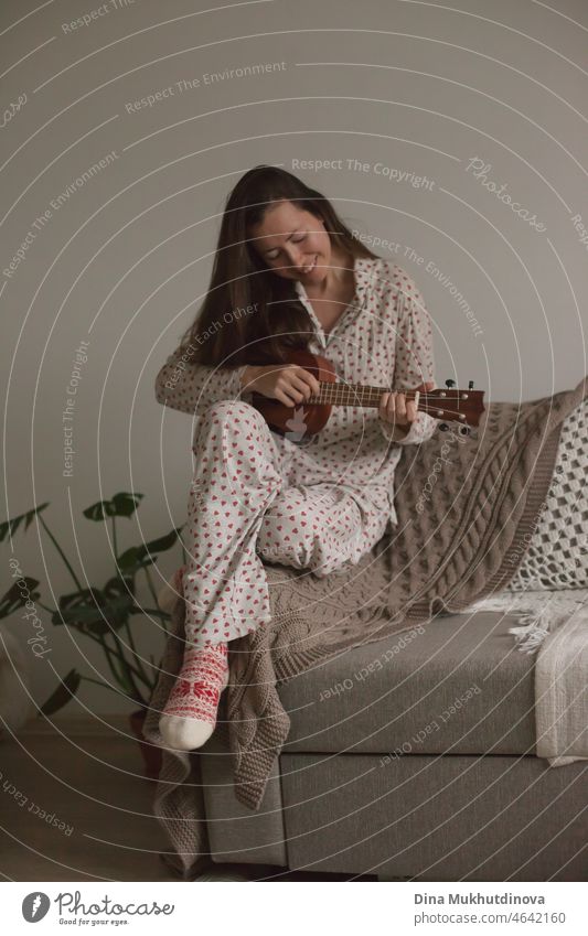 Young woman at home in pajamas playing guitar sitting on a couch or sofa. Millennial female musician in cozy apartment playing ukulele dressed in holiday christmas season pajamas. Talented young person practicing playing ukulele.