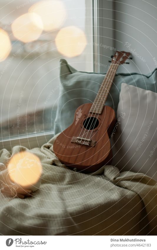 Ukulele at home on the windowsill with cozy blankets and pillows on a cold rainy day. Cozy mood spending time at home. Melody and music hobby - small guitar for playing music.