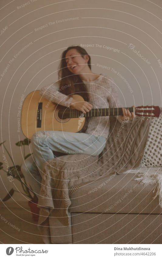 Happy young woman playing guitar and singing, sitting on a couch in cozy room at home in modern apartment. Music and hobby at home or at party with friends.