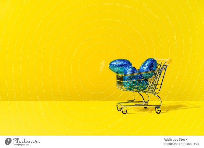 A shopping cart with colourful blue foil eggs on a yellow background festive purchase sale store copy space market business basket celebration concept