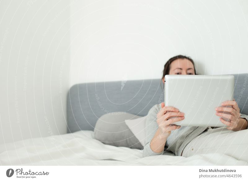 Young woman working at home office lying with tablet on the bed. Watching movies or working online. Informal home office. Gray color palette image of person using technology mobile  device.