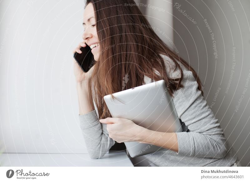 Young millennial woman with brown hair talking on the mobile phone and holding tablet device. Gray color palette. Woman working in office or at home holdng mobile devices. Communicationa at work and online business concept.