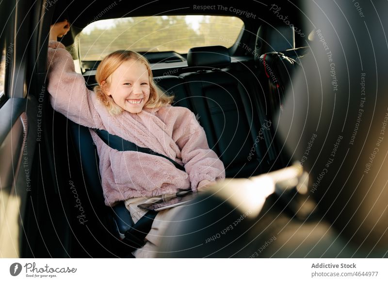 Cheerful girl sitting in car kid automobile passenger backseat ride trip commute having fun tablet modern happy device vehicle positive cheerful smile journey