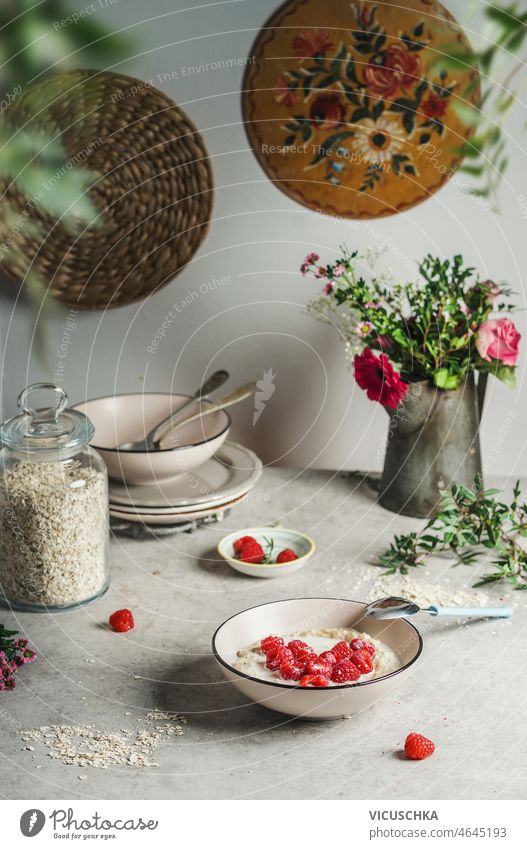 Bowl with oats porridge and raspberries heart on grey table with kitchen utensils flower bunch stapled bowls healthy homemade breakfast at home front view