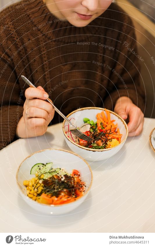 Woman in brown sweater eating a poke bowl or salad at the restaurant. Colorful healthy food - vegetables and fish and seafood. hawaiian overhead white dining