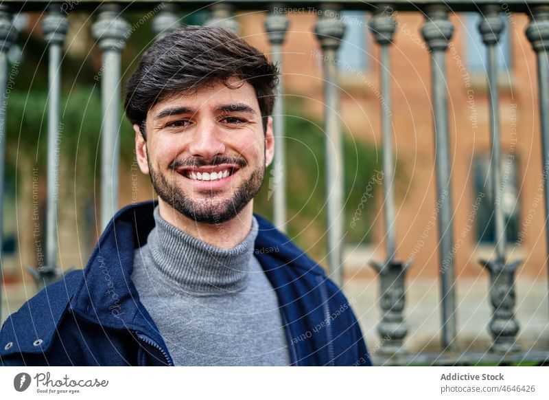 Cheerful man standing near fence and looking at camera individuality confident masculine cool appearance gaze urban park beard turtleneck self assured