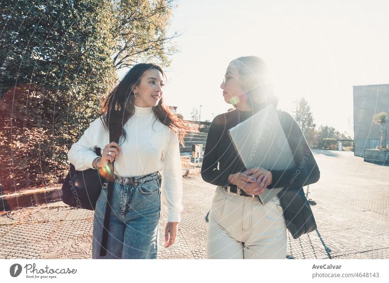 Two young student woman going to class while talking and laughing outdoors the faculty during a bright day. University studies, college papers, modern style. Preparing a presentation for class.