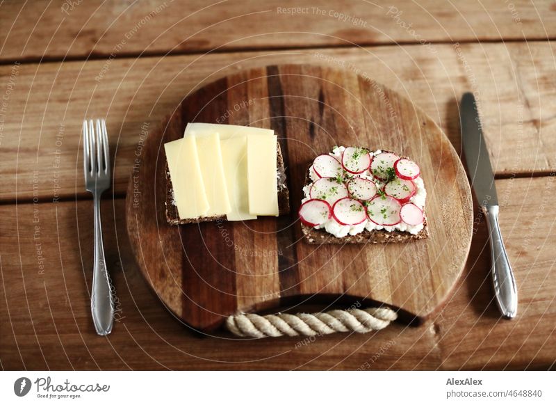 Healthy breakfast - one slice of brown bread topped with Gruyère and one with grainy cream cheese, radishes and cress - on a wooden breakfast board with cutlery on the wooden table