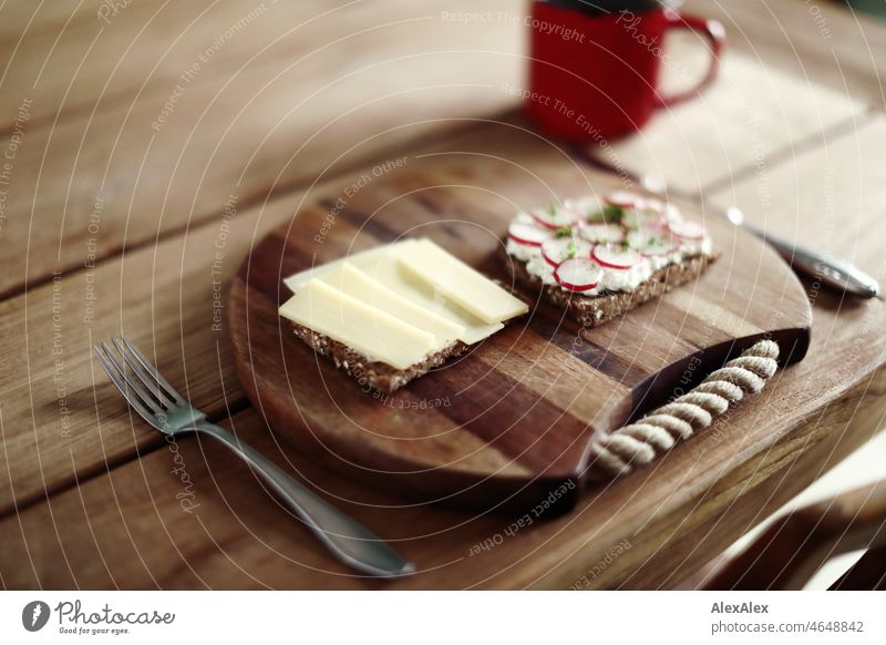 Healthy breakfast - a slice of brown bread topped with Gruyère and one with grainy cream cheese, radishes and cress - on a wooden breakfast board with cutlery on the wooden table with coffee in a red cup