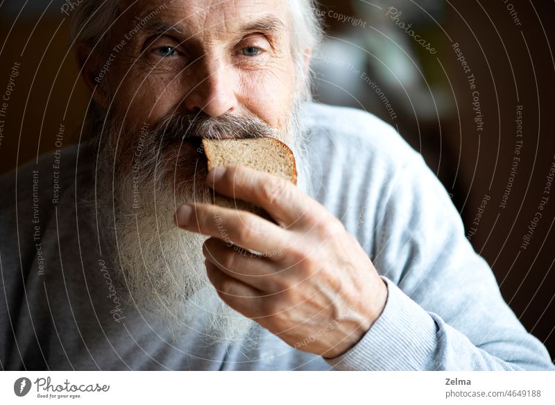 Old sad man with a long gray beard sitting by the table and eating soup and bread old senior elderly holding bred closeup face wrinkles eyes moustaches hand