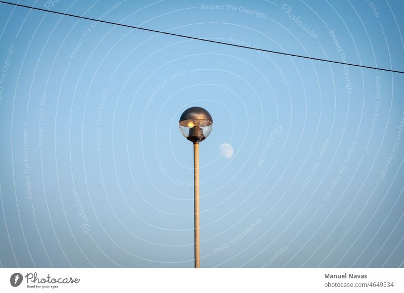 Light pole at sunset in front of a blue sky with the moon, the pole is hit by the last rays of sunlight of the day, there is also an electric cable. copy space