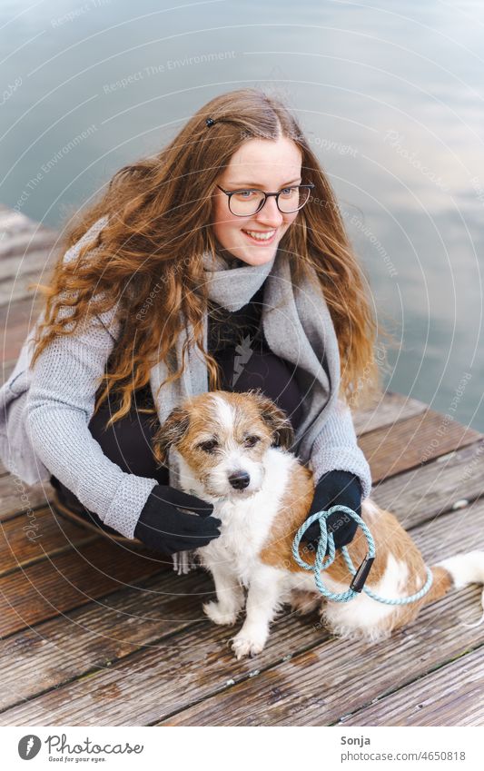 A young woman with glasses and long hair sits with a small dog on a wooden walkway. Woman youthful Dog Terrier Small Pet Animal Cute Exterior shot Lifestyle