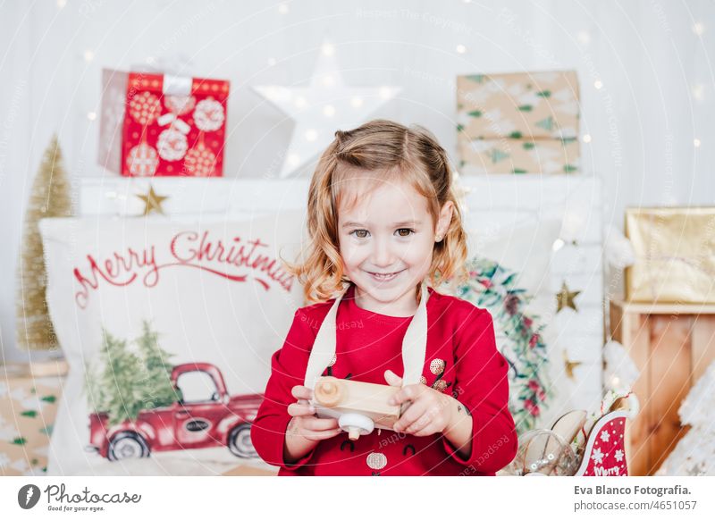 happy little girl at home holding toy camera wearing red christmas dress at home over christmas decoration. Holiday concept kid smiling caucasian blonde