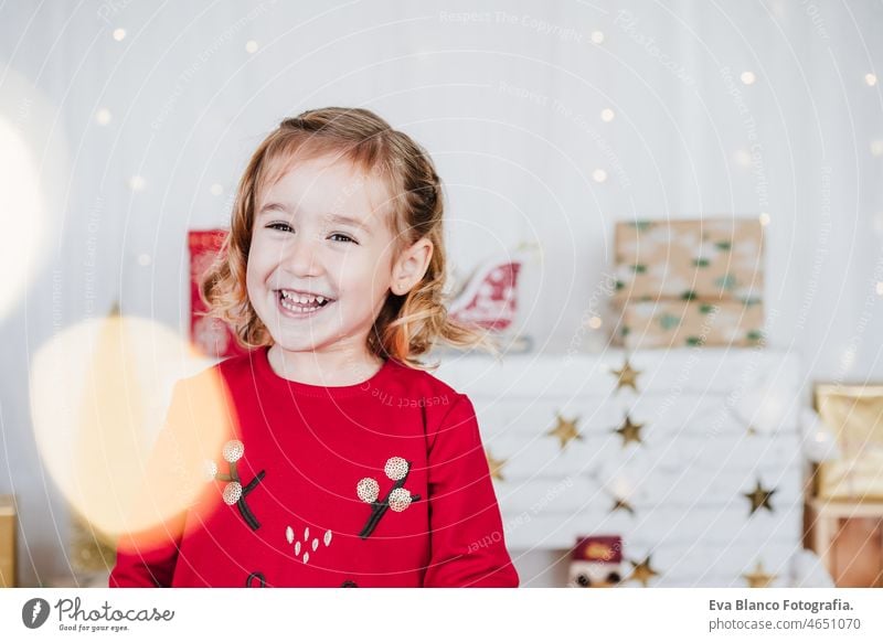portrait of happy little girl at home wearing red christmas dress at home over christmas decoration. Holiday concept kid smiling caucasian blonde 3 year old