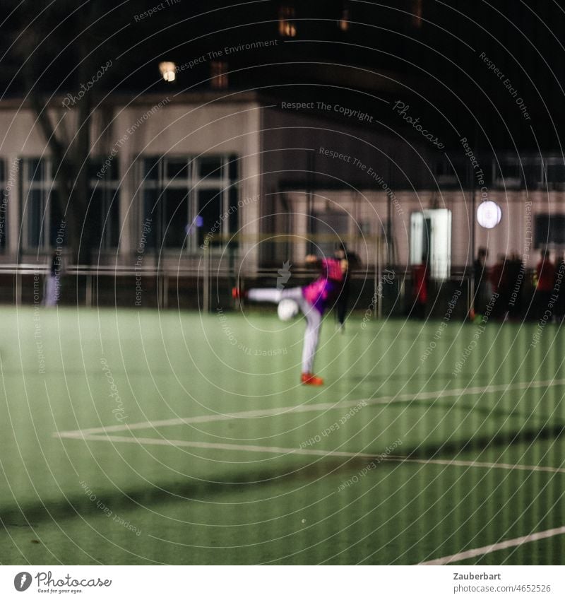 Soccer player shooting goal, shadowy at night on illuminated sports field through fence Foot ball soccer player Ball Shot at goal Movement Sporting grounds