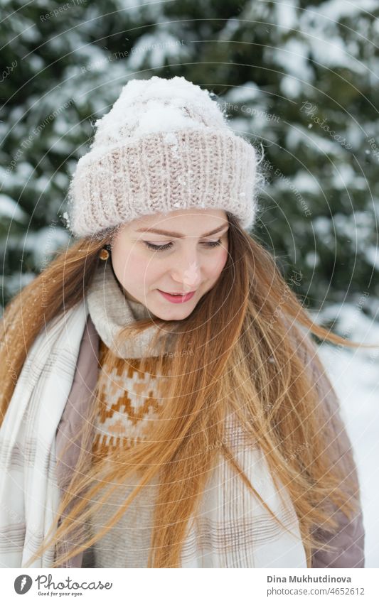 Beautiful caucasian woman with long hair walking in winter forest with snow on her knitted beanie hat. Winter fashion and stylish outfit. Real people having fun in winter, enjoying fresh air in nature with snowy fir trees.