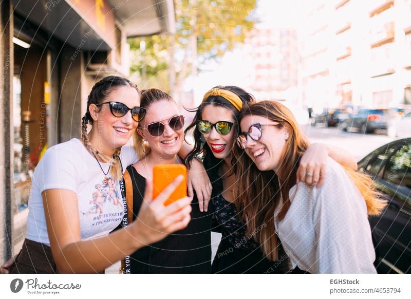 Group of 4 young women friends enjoying a sunny day in the city, posing for a picture looking straight ahead carefree cheerful city life enjoyment fashion