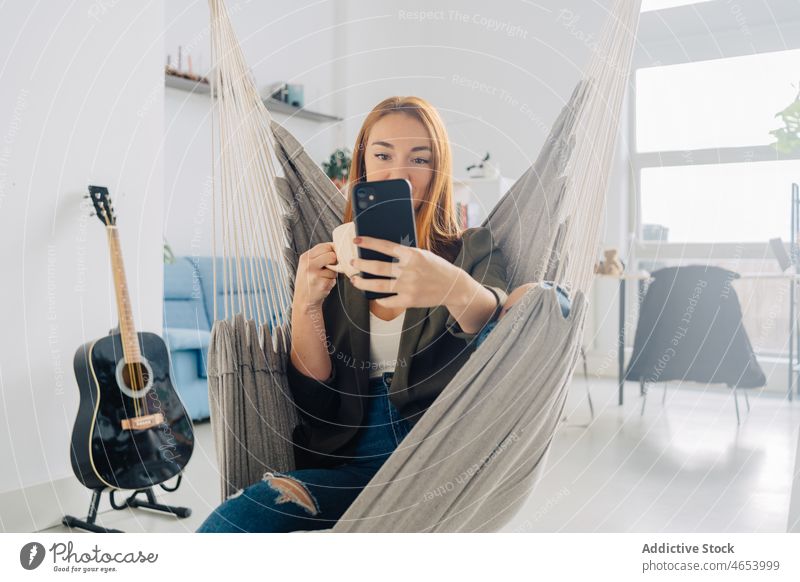 Woman taking selfie in hammock with coffee woman smartphone using self portrait chill relax free time female hot drink at home rest red hair pastime cellphone