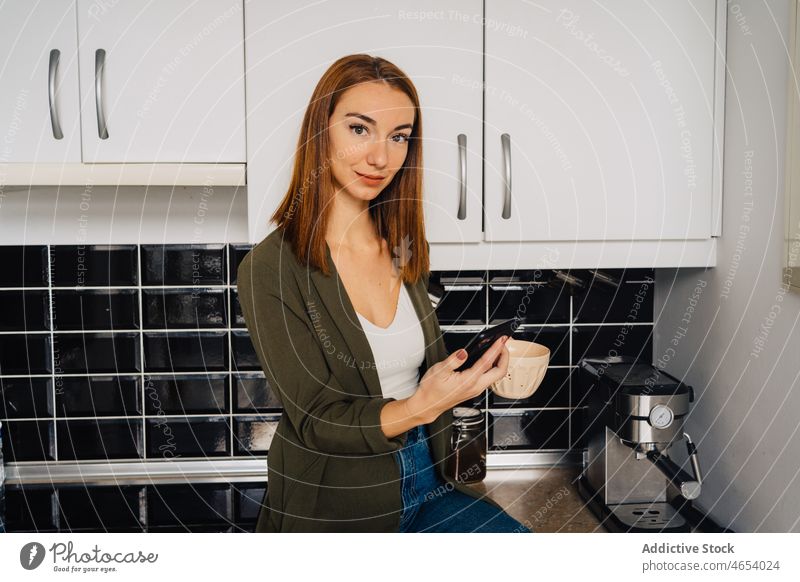 Female with cup of hot drink using cellphone in kitchen woman smartphone counter coffee texting female concentrate sit denim ripped rest mobile tea sms lounge