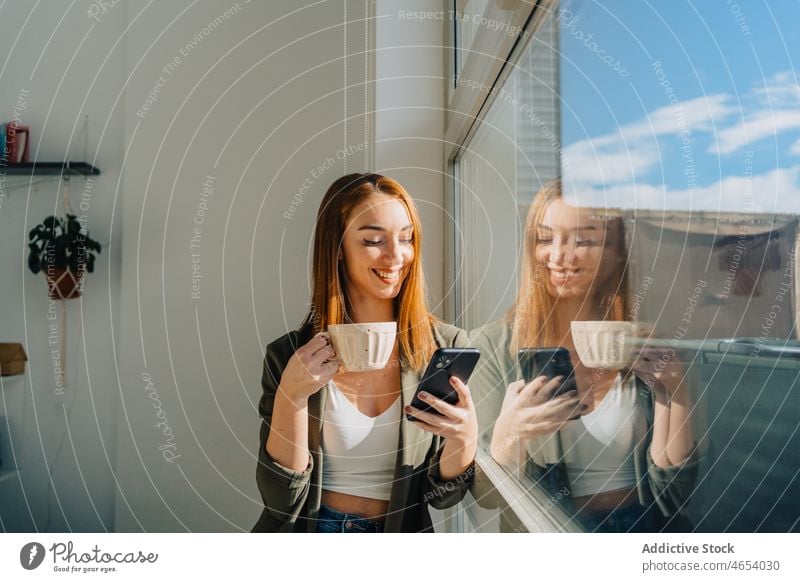 Cheerful lady with cup of coffee browsing smartphone near window woman cellphone cheerful using drink hot drink smile reflection comfort female rest texting