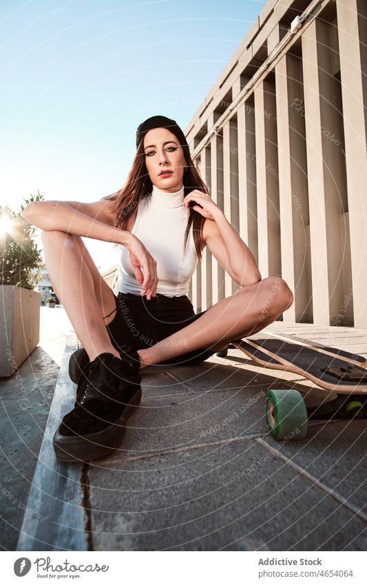 Young woman sitting on longboard in park style city urban summer activity appearance allure female slim young charming striped wall brunette concrete hobby