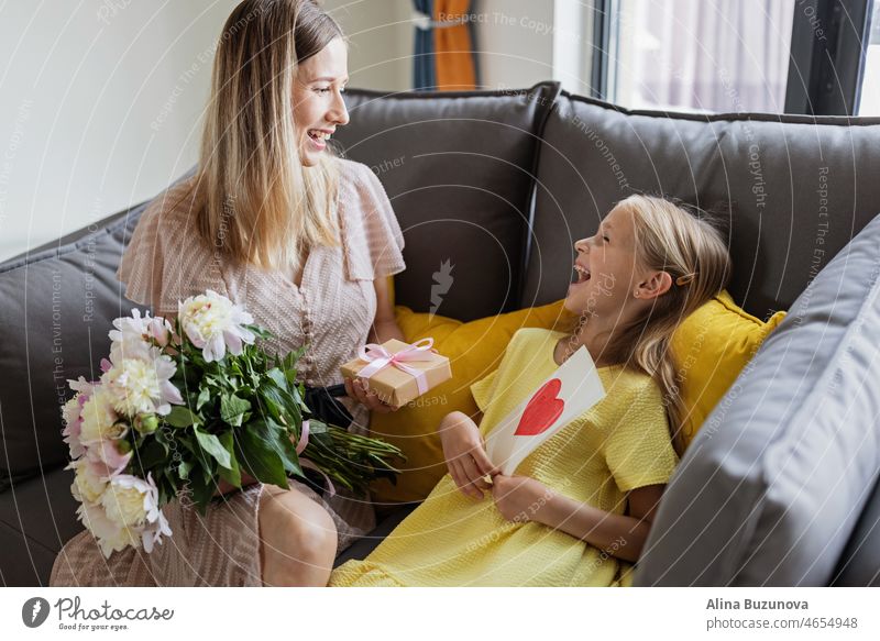 Child daughter congratulates mother and gives present card, gift and bouquet of flowers at home. Mom and girl smiling and hugging on couch. Happy mothers day celebration