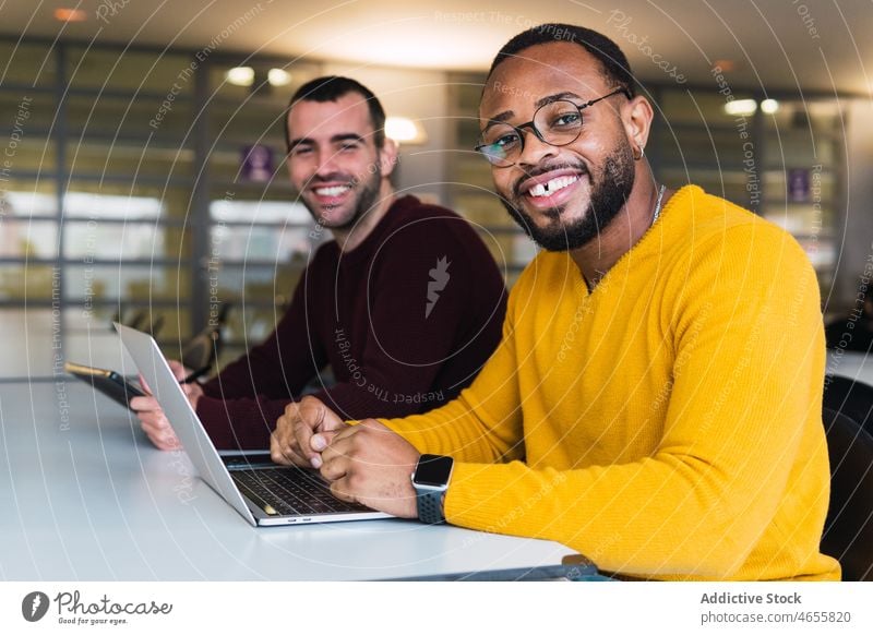 Positive diverse guys with laptop and tablet in modern workspace men colleague partner coworker collaborate teamwork workplace freelance using male cheerful