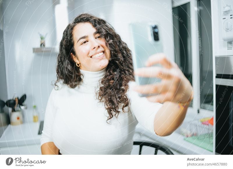 Young plus size spanish woman blogging about his diet and eating habits at a modern kitchen taking pictures and cooking. Sitting on a modern stool and having fun. Losing wight concept