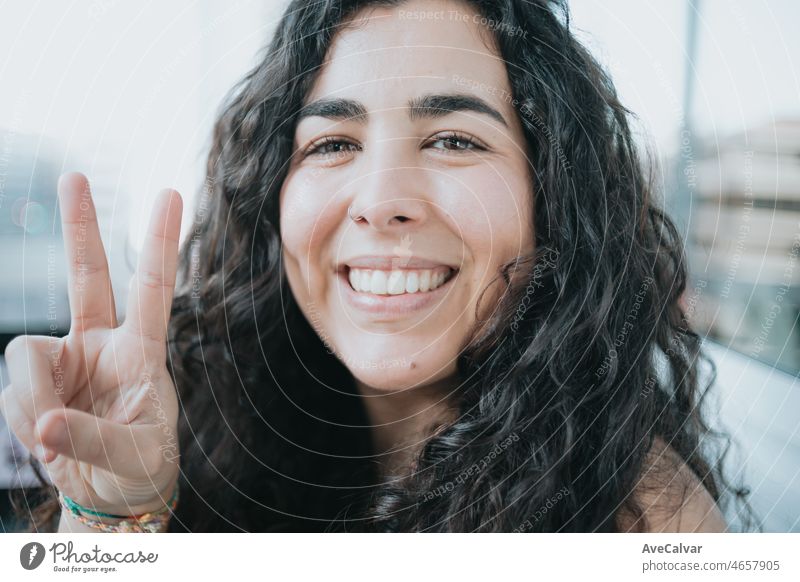 Young spanish woman with curly hair showing victory sign with a cute smiling face while working on his laptop. Close up portrait. Freelancer happy to work by her own