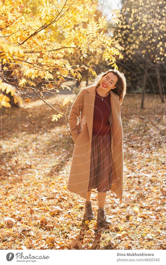 Beautiful woman in beige coat, burgundy sweater, brown skirt and red lipstick make up, walking in autumn park. Portrait of a girl in autumn nature with yellow foliage.
