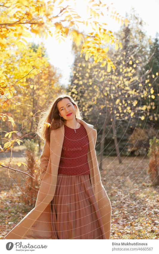 Beautiful woman in beige coat, burgundy sweater, brown skirt and red lipstick make up, standing in autumn park. Portrait of a girl in autumn nature with yellow foliage.