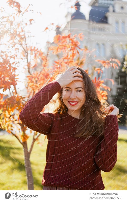Young woman in burgundy sweater and red lipstick make up, standing in autumn park near historic building. Portrait of a girl in autumn park with orange red foliage.