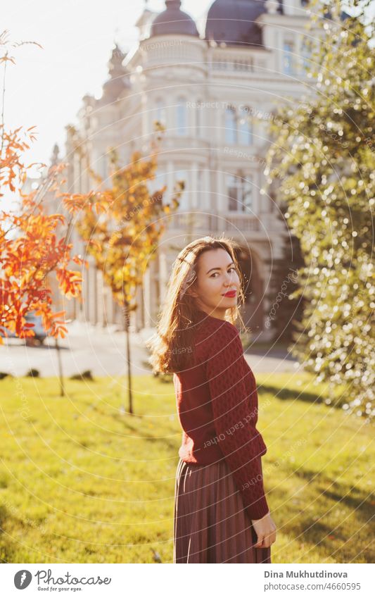 Young woman in burgundy sweater, brown skirt and red lipstick make up, standing in autumn park near historic building. Portrait of a girl in autumn park with orange red foliage.