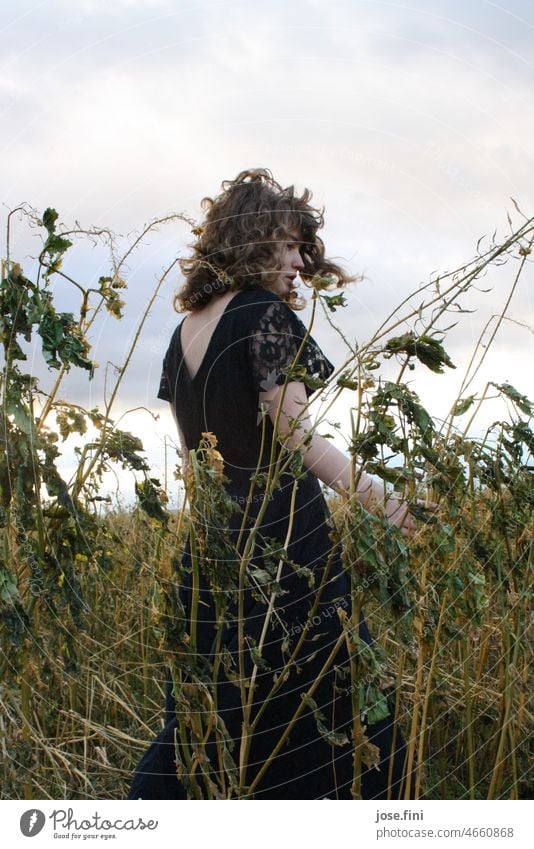 Young woman with curls stands in dark dress in field, dried grasses around her. Plant naturally Exterior shot Full-length Day Girl Nature Meadow Field Slim