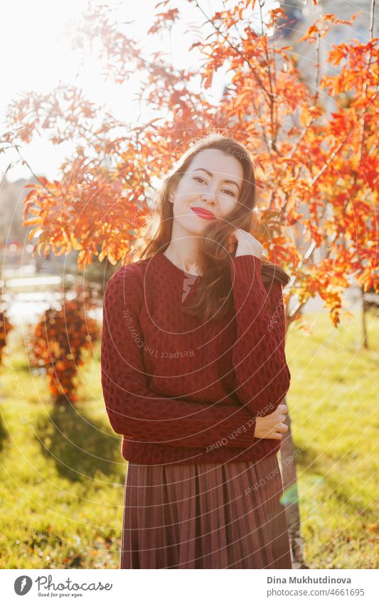 Young woman in burgundy sweater, brown skirt and red lipstick make up, standing near autumn tree. Portrait of a girl in autumn park with orange red foliage.