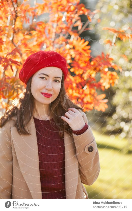 Young woman in red beret, beige coat and burgundy sweater and red lipstick make up, standing near autumn tree. Portrait of a girl in autumn park with orange red foliage.