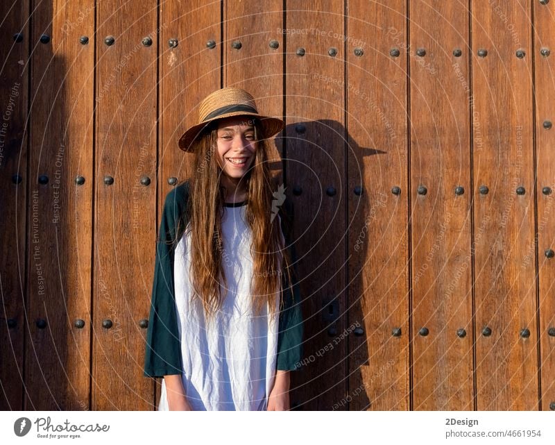 Young happy teenager female with summer clothes and hat standing against old wooden outdoors door while looking at camera girl smiling toothy long hair outside