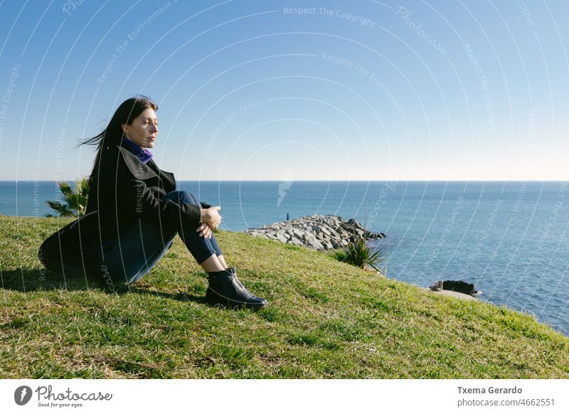 Middle-aged woman meditating in front of the sea on a winter day 45-50 years beach grass mature sun wind lonely emotion outside calm copy space pretty