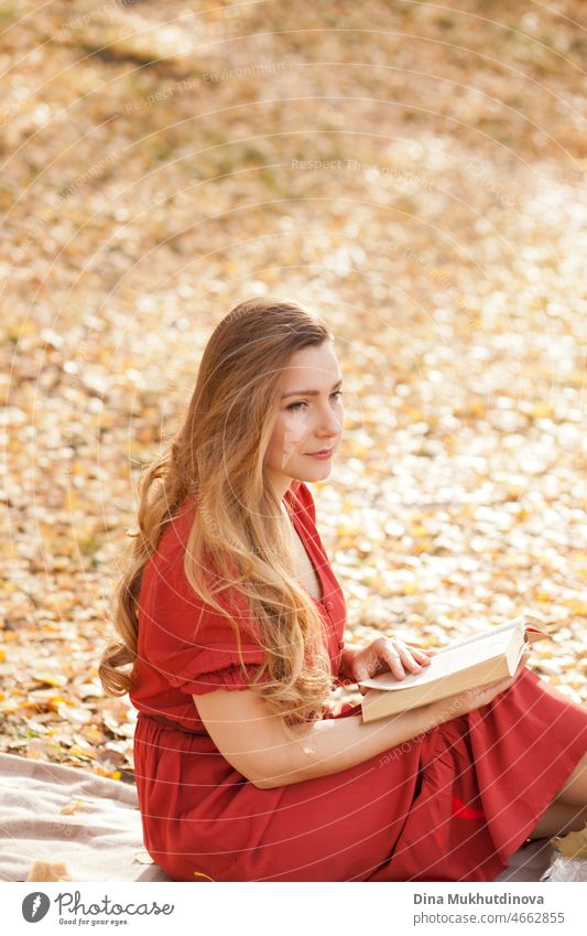 Beautiful woman with long hair in red dress reading a book in autumn park outdoors in nature. Romantic book lover reading outside on a sunny autumn day.